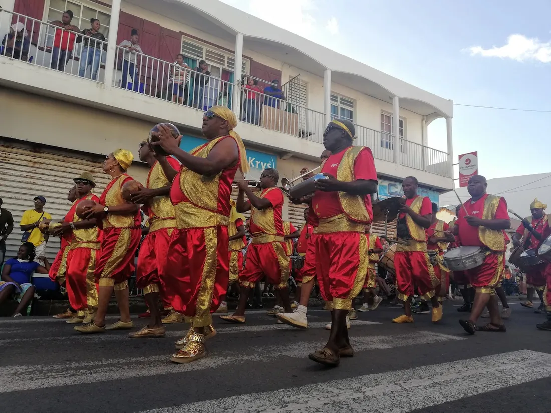 Groupe a po de percussionnistes en rouge et or defilant dans une rue bordee de batiments avec spectateurs aux balcons en Guadeloupe