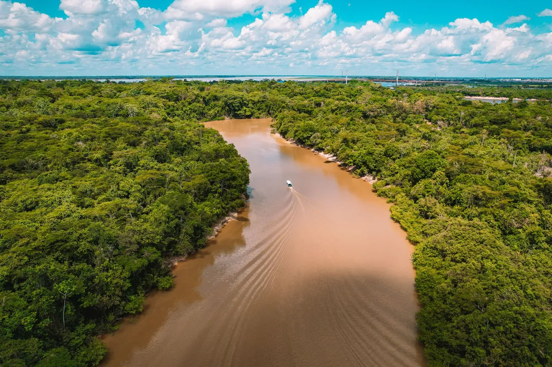 Vue aerienne d'une riviere sinueuse aux eaux ocre traversant la foret amazonienne dense de Guyane, avec une petite embarcation