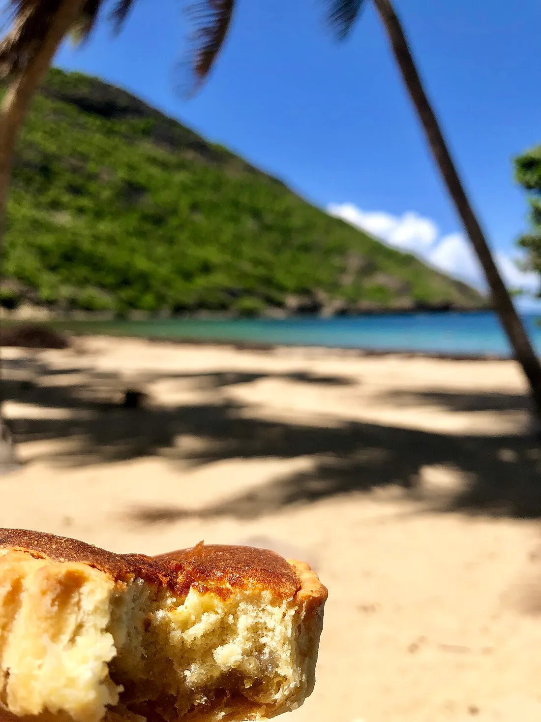 Une part de tourment d'amour, la pâtisserie traditionnelle des Saintes, tenue à la main devant une plage de l'archipel en Guadeloupe